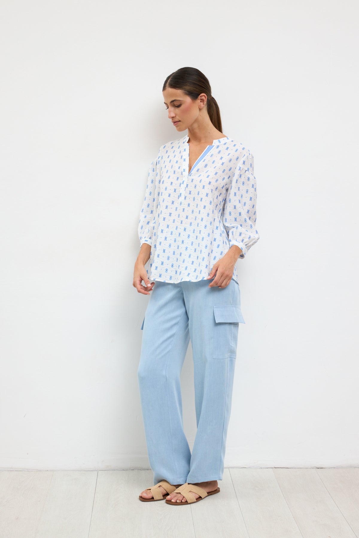 Woman wearing a white blouse with blue patterns and light blue pants against a white background